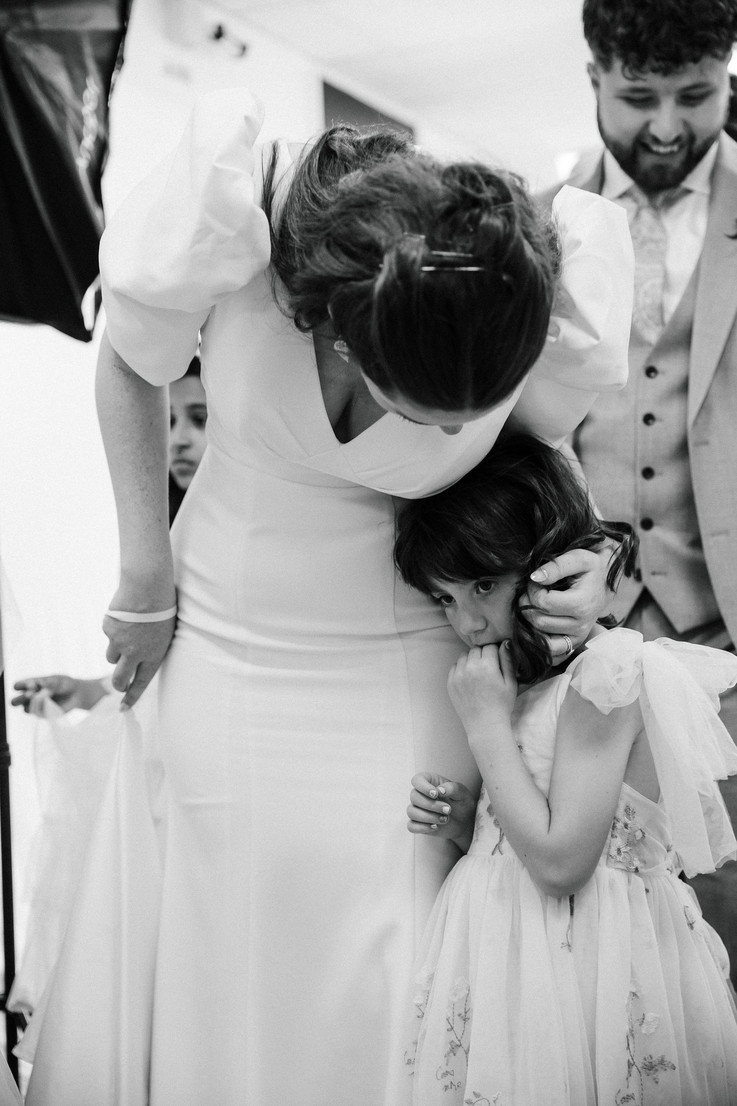 Bride tenderly embracing young flower girl in white dress at wedding reception in black and white photo.