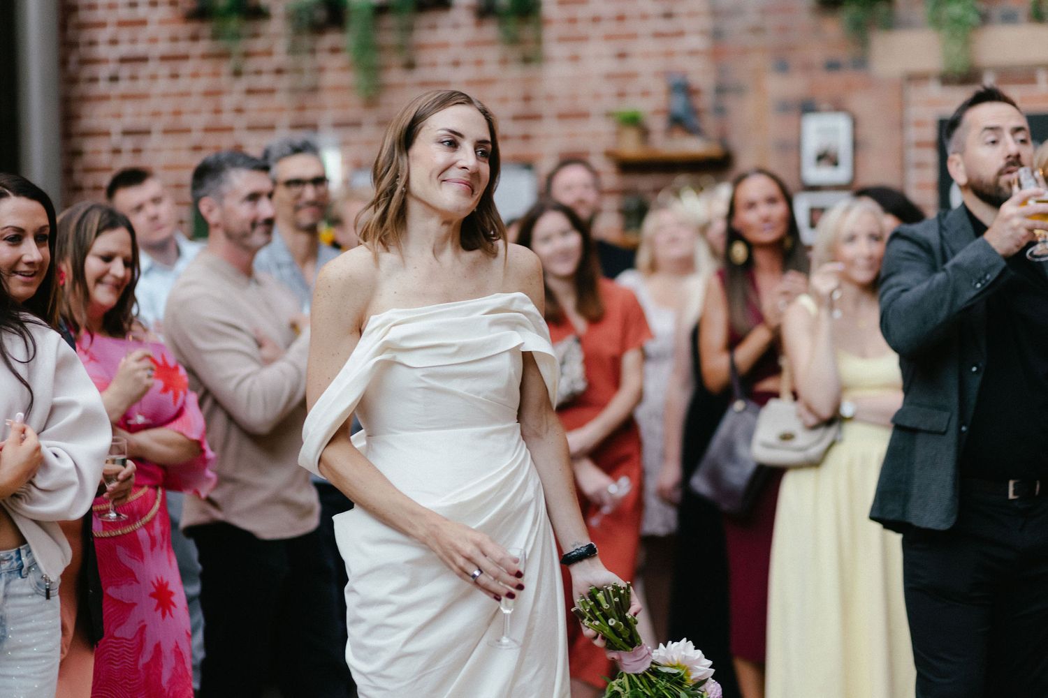 Bride in white off-shoulder gown smiling at wedding reception with guests gathered in brick-walled venue.