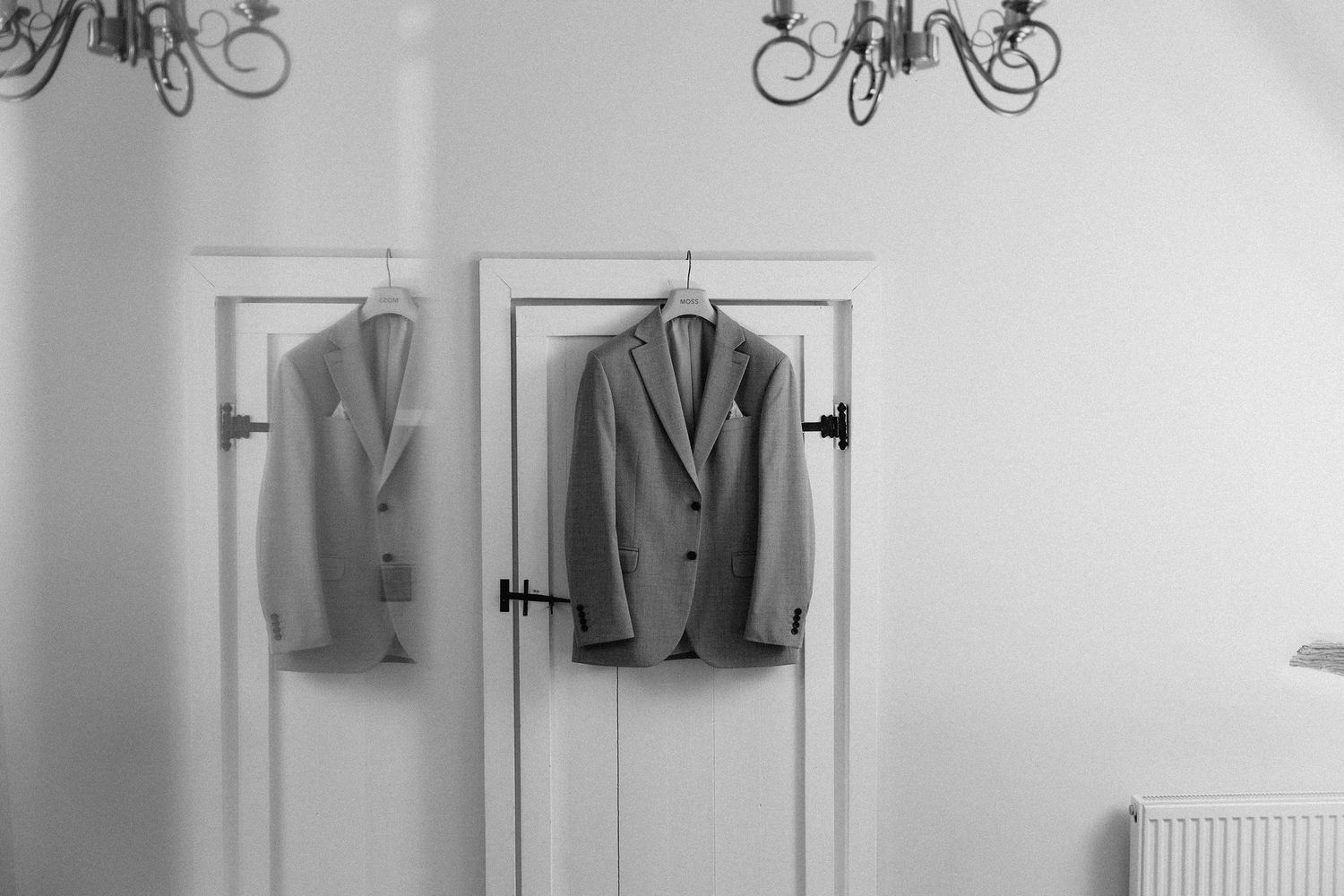 Two elegant suits hanging on doors in a bright white room with chandelier lighting in black and white photo.