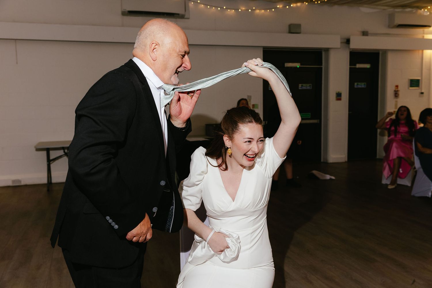 Bride in white gown laughing while pulling a man's tie on a wedding reception dance floor.