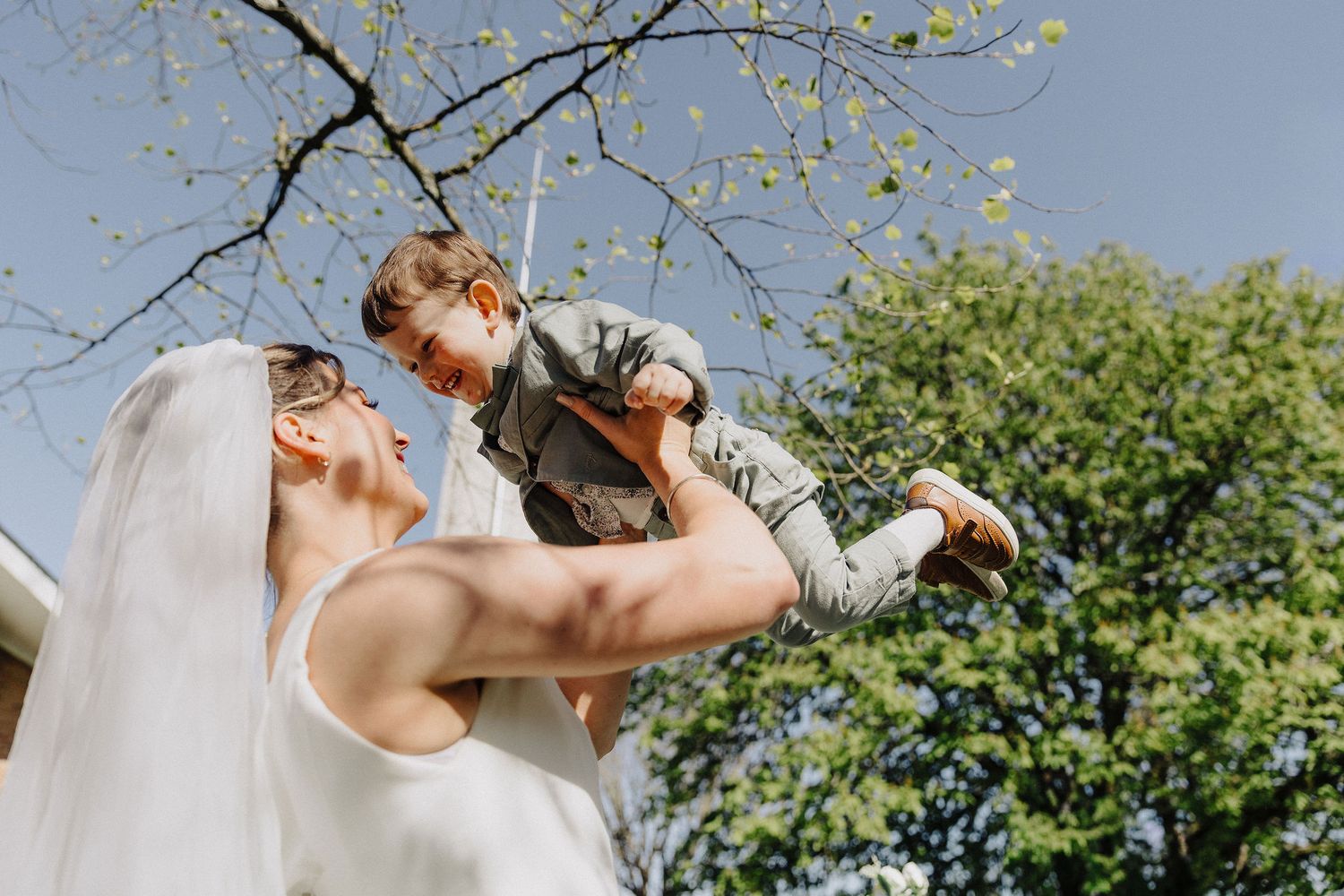 Bride in white dress and veil lifting a young child in the air outdoors on a sunny wedding day.