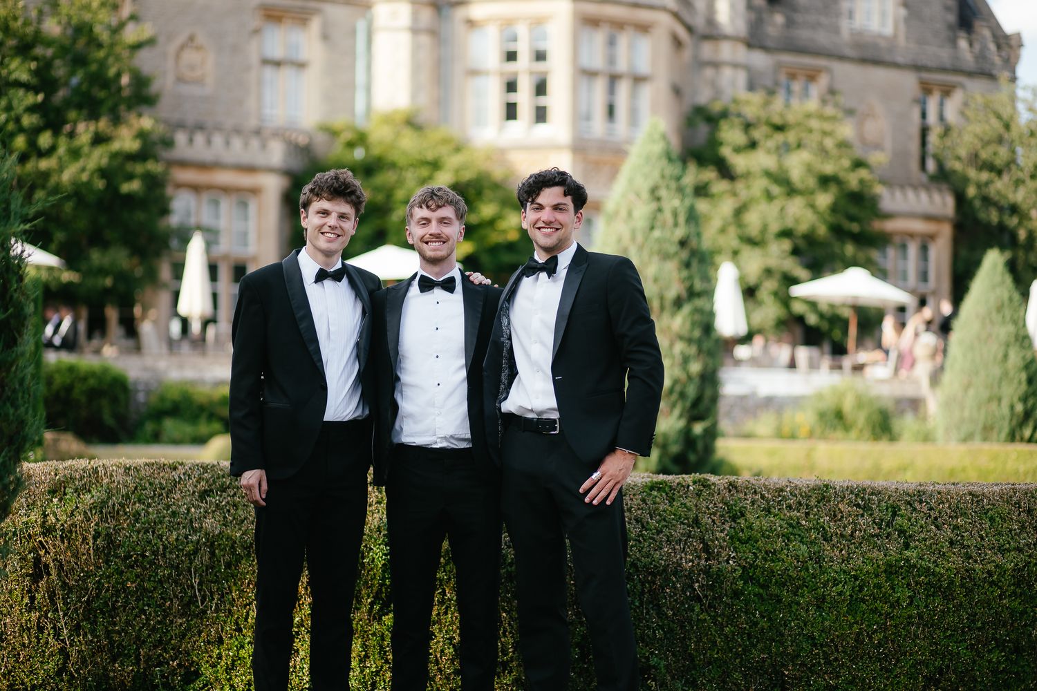 Groomsmen in tuxedos pose for formal photos in front of historic building.