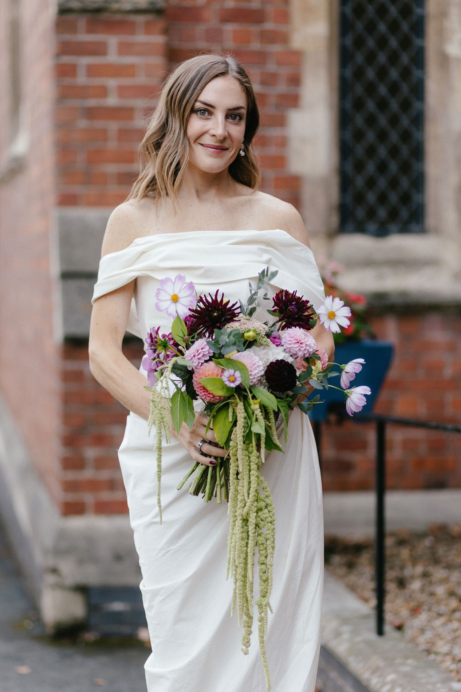 Bride in off-shoulder white gown holding a lush bouquet of dahlias and wildflowers, posing before a brick building.