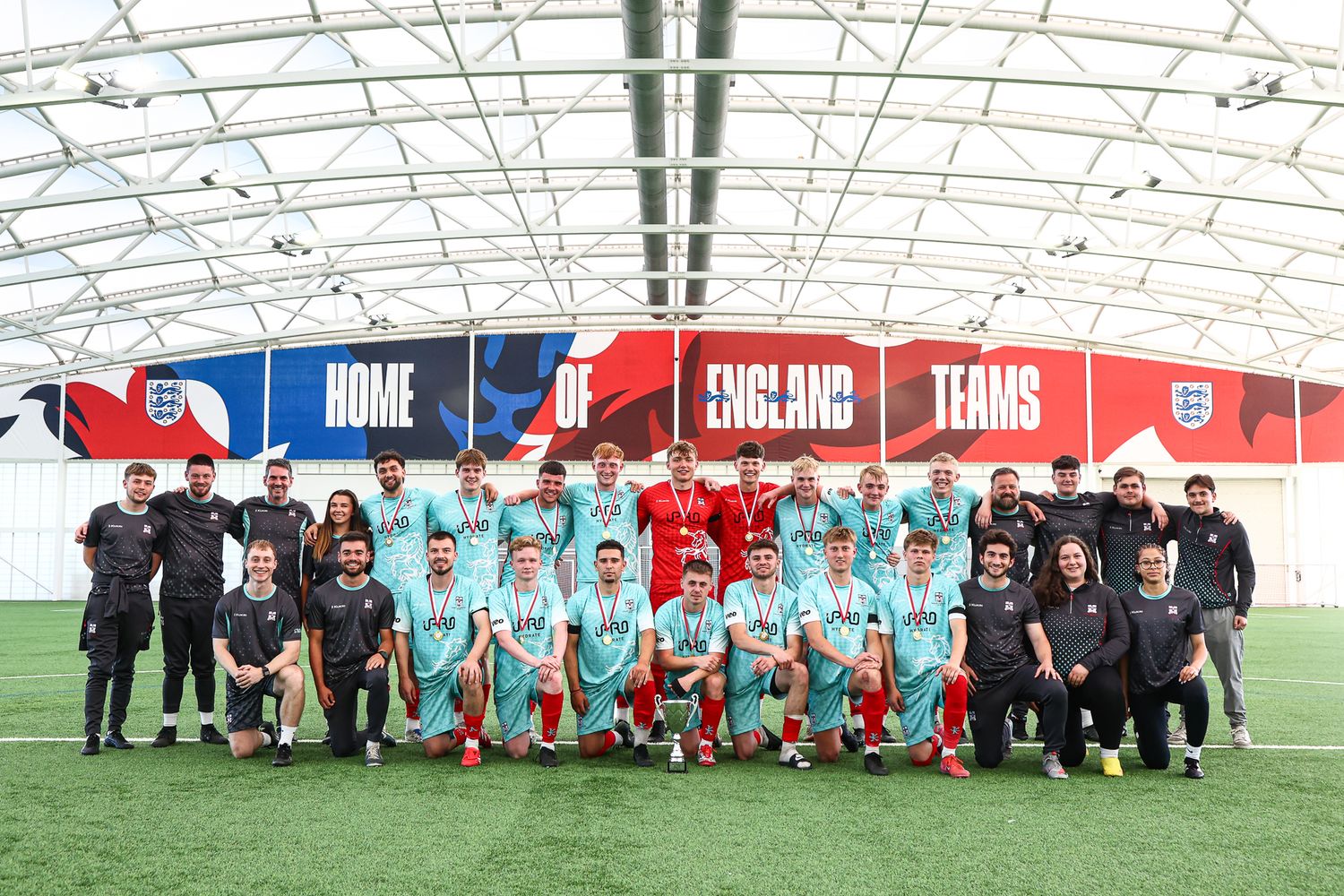 Wide angle team photo showing soccer players and coaching staff on indoor training pitch.