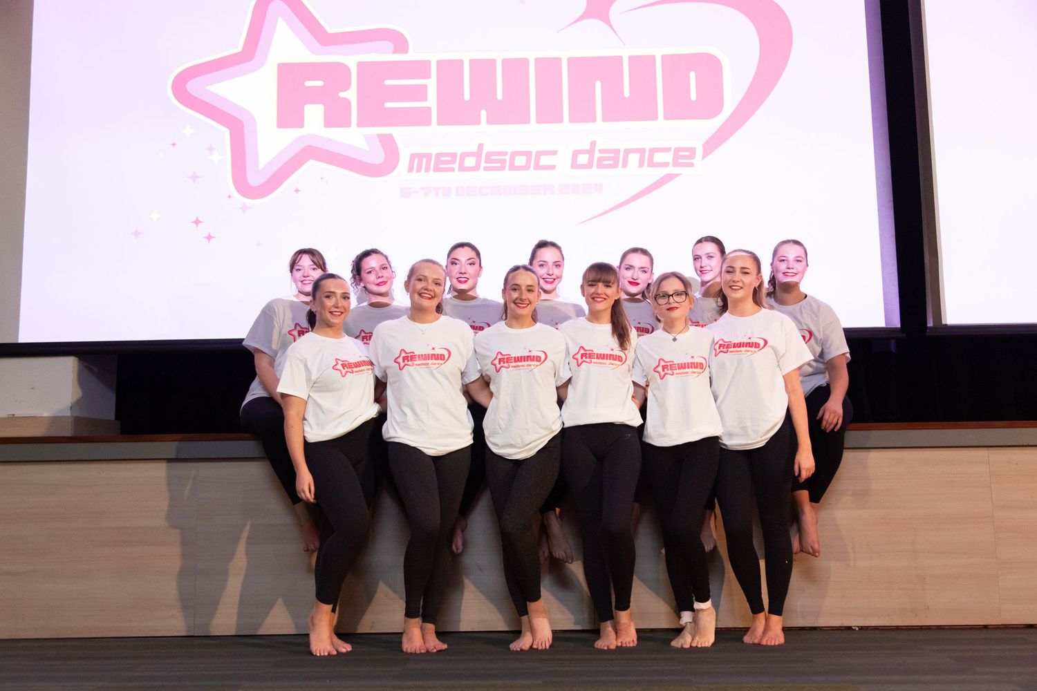 Dance group in matching white shirts poses together on stage with pink Rewind logo backdrop.