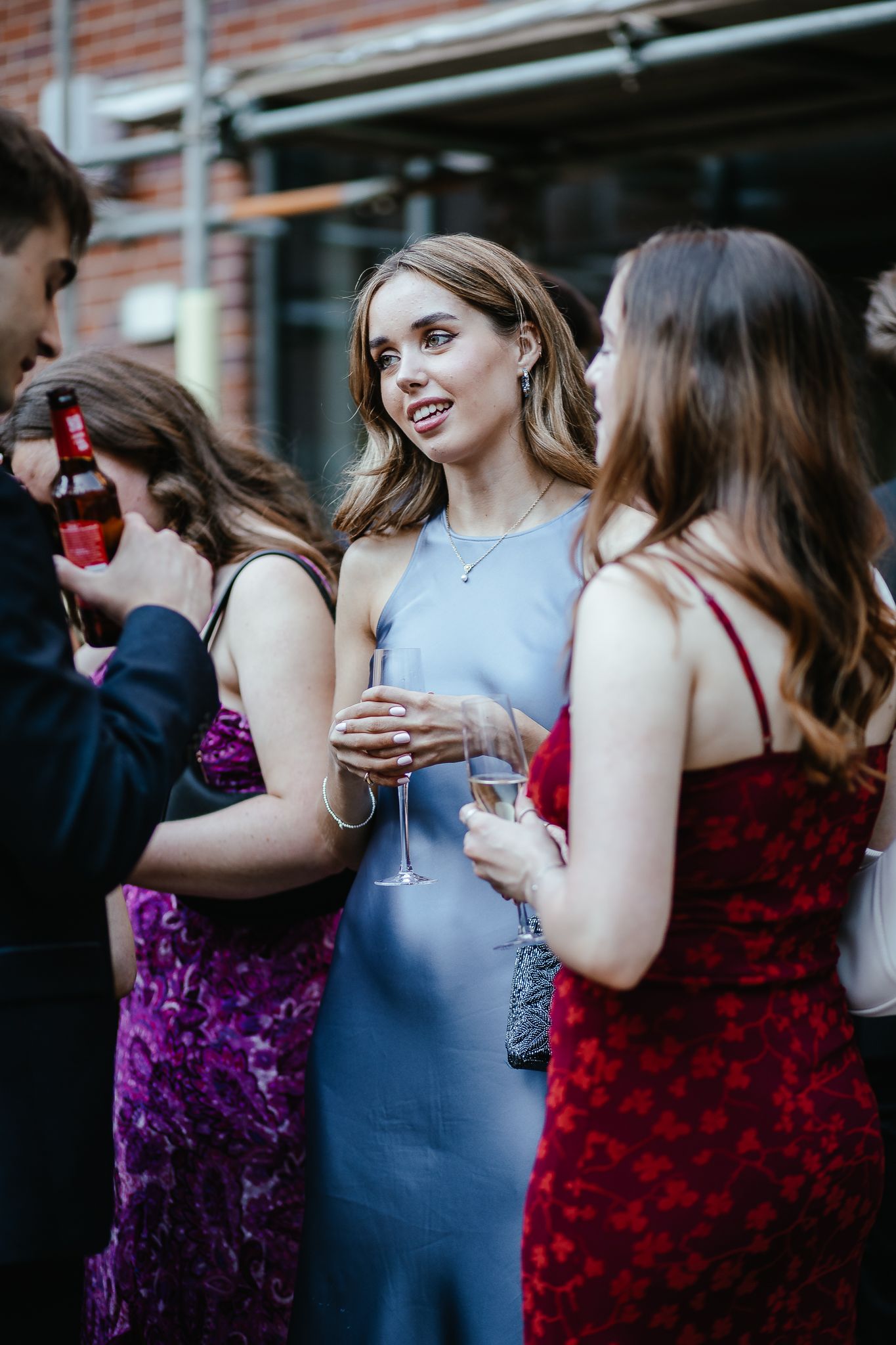 Wedding party members in jewel-toned dresses holding champagne glasses against brick wall.
