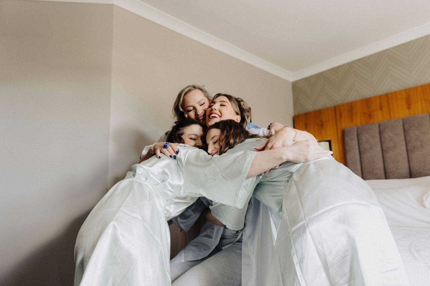 Group of bridesmaids sharing a joyful hug in a hotel bedroom while wearing white robes before a wedding.