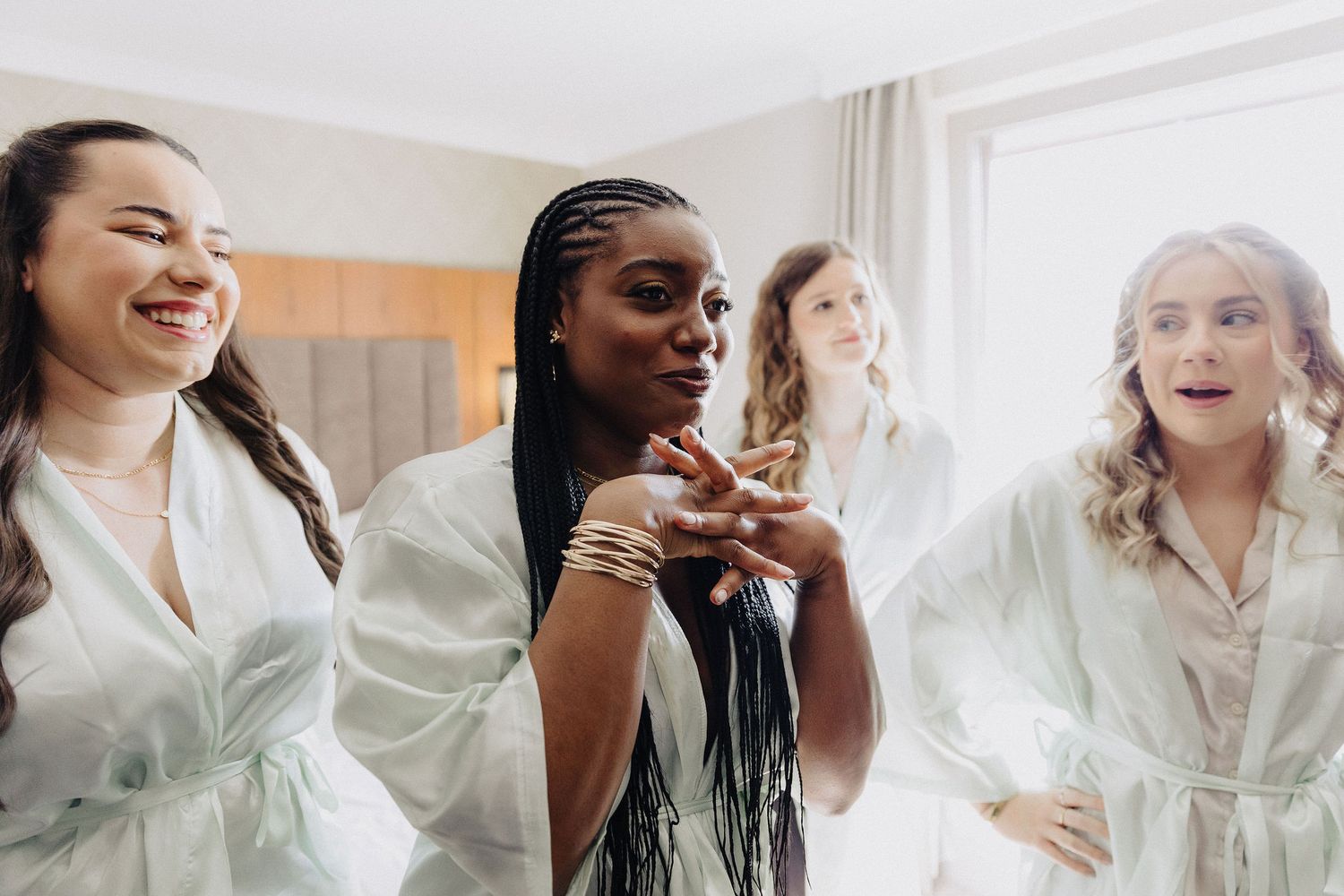 Four bridesmaids in white robes laughing and getting ready together in a bright hotel room before a wedding.