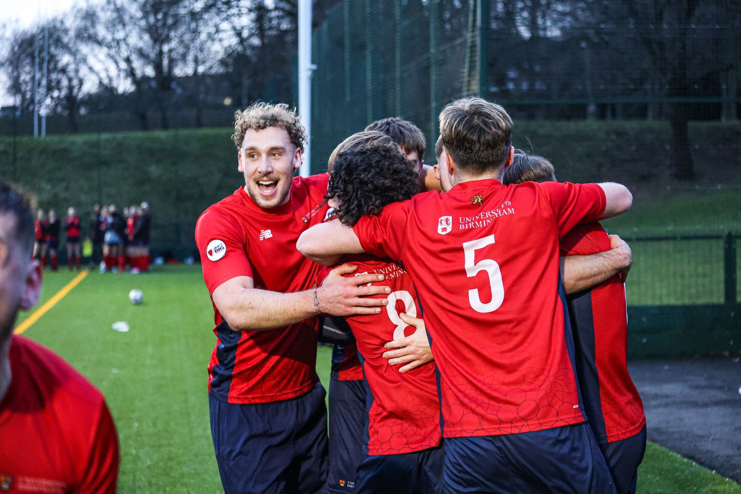 Soccer teammates celebrating with group hugs on the field wearing red jerseys.