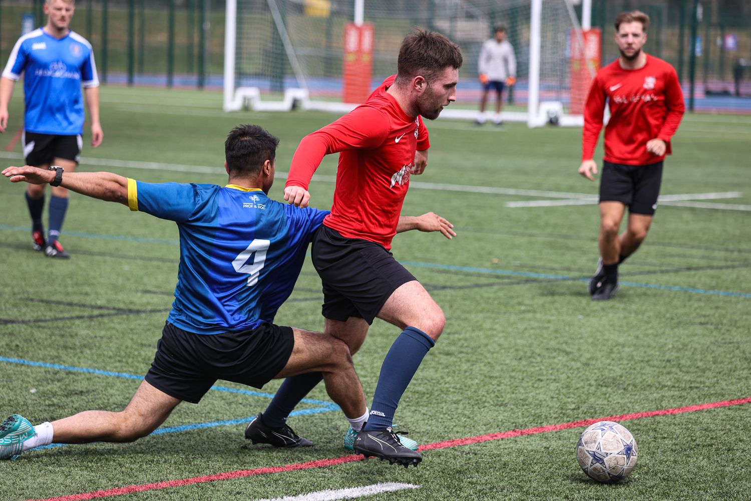 Soccer players in red and blue jerseys competing for possession of the ball on a turf field.