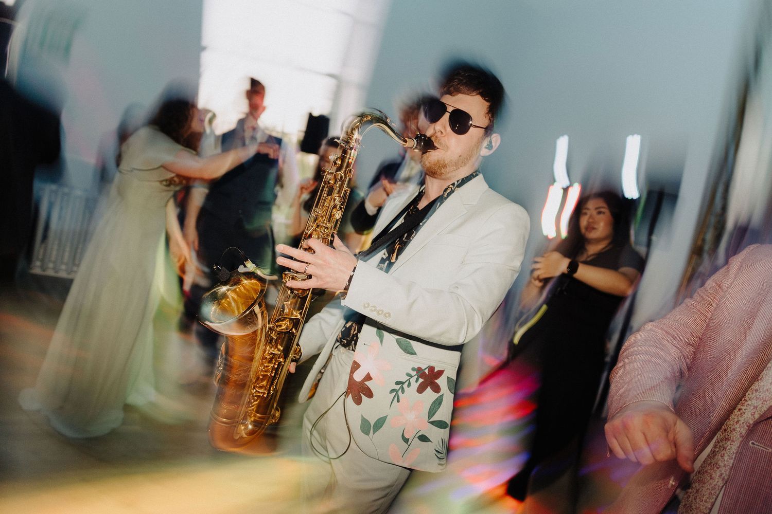 Saxophonist in floral jacket and sunglasses performs at lively wedding reception dance floor party.