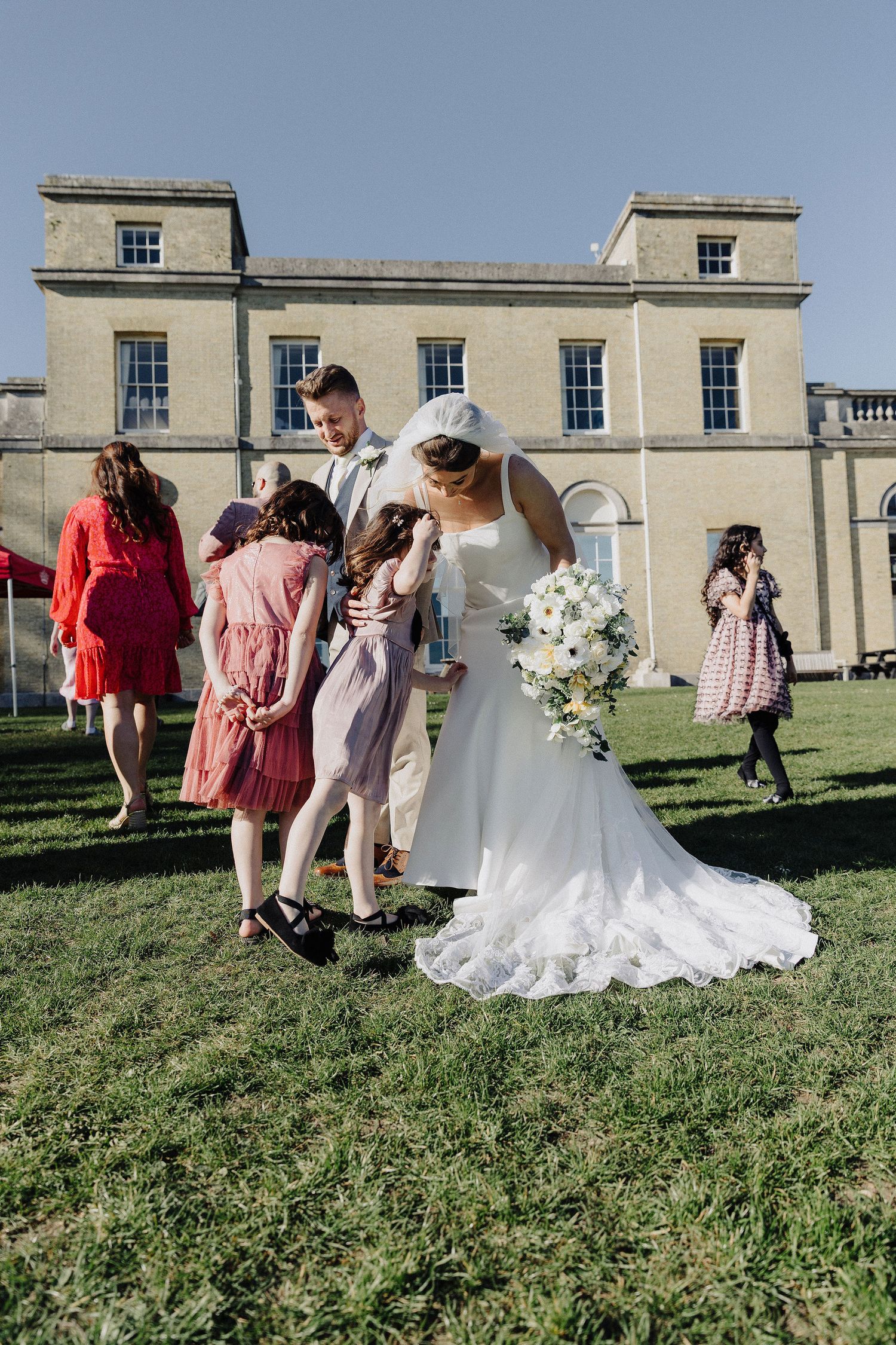 Bride and groom interact with young flower girls on lawn outside elegant stone manor house wedding venue.