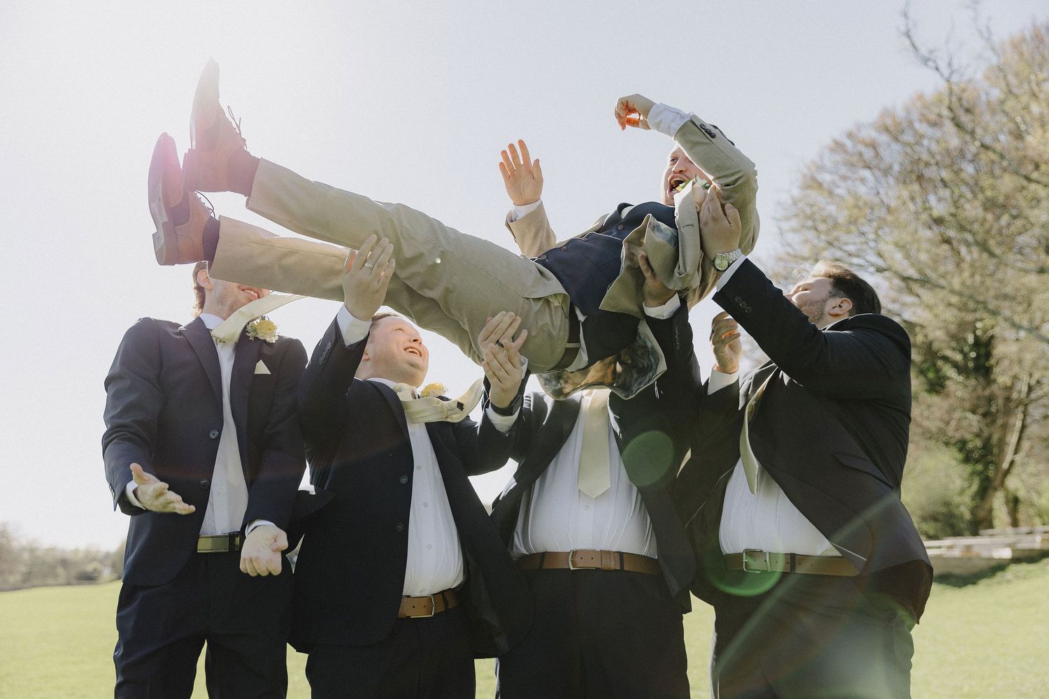 Groomsmen joyfully tossing the groom into the air outdoors on a sunny wedding day.