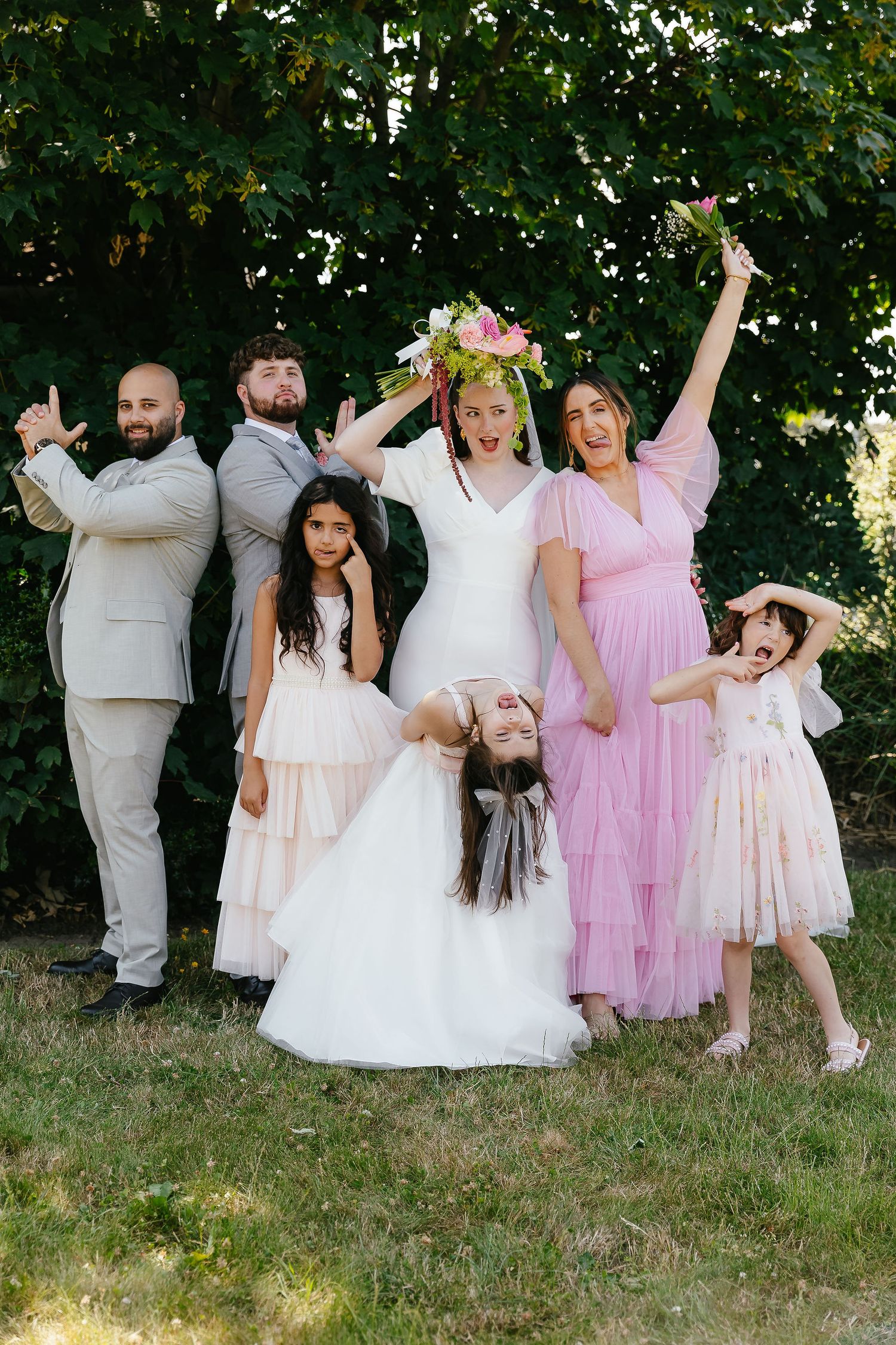 Wedding party posing playfully outdoors with bride in floral crown, bridesmaid in pink, groomsmen, and children.