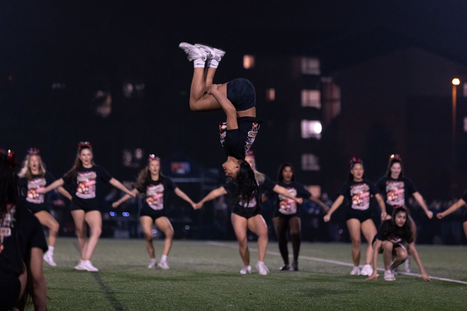 Cheerleaders performing a dramatic nighttime stunt with someone airborne under stadium lights.