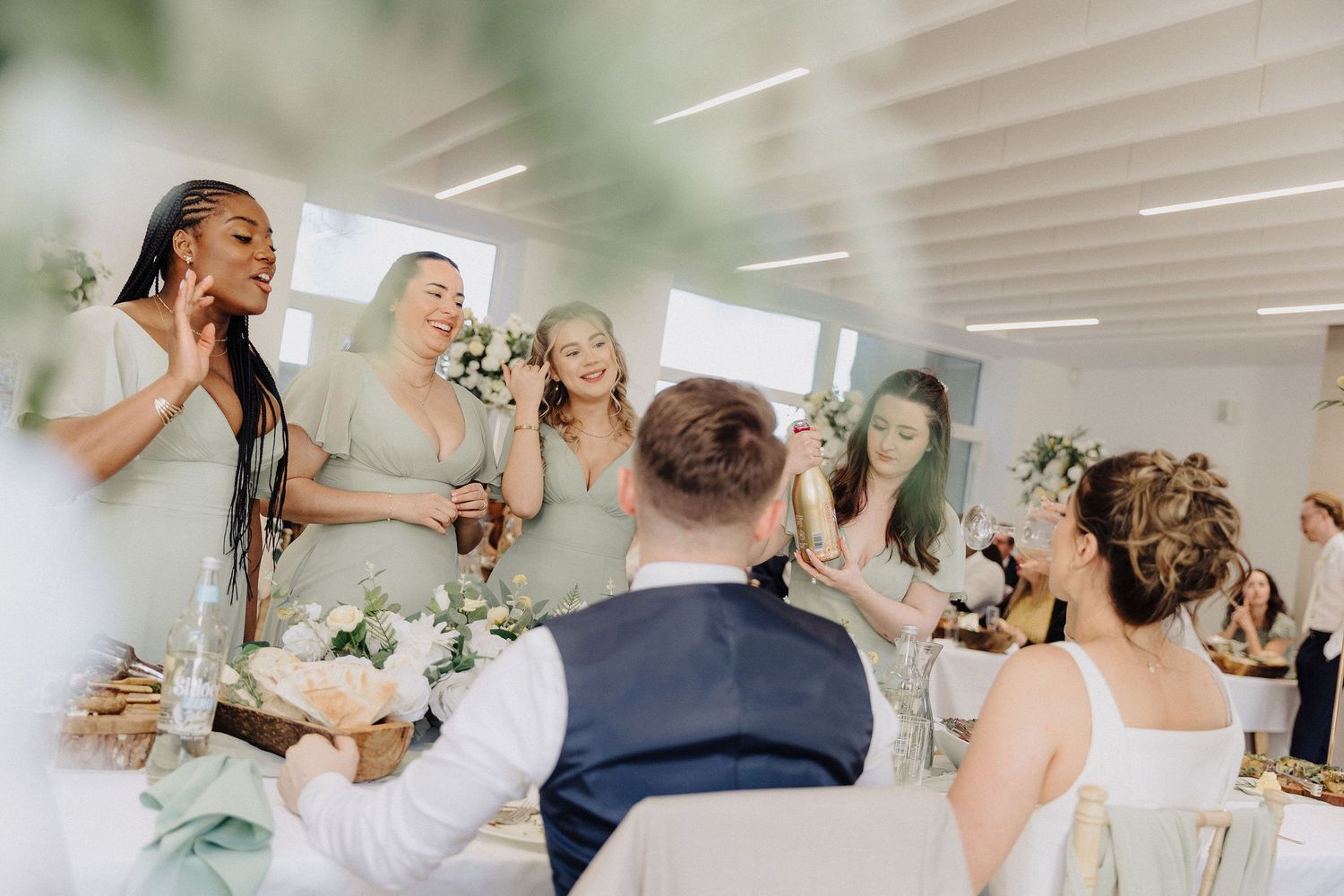 Bridesmaids in sage green dresses laughing and celebrating with bride at elegant wedding reception.