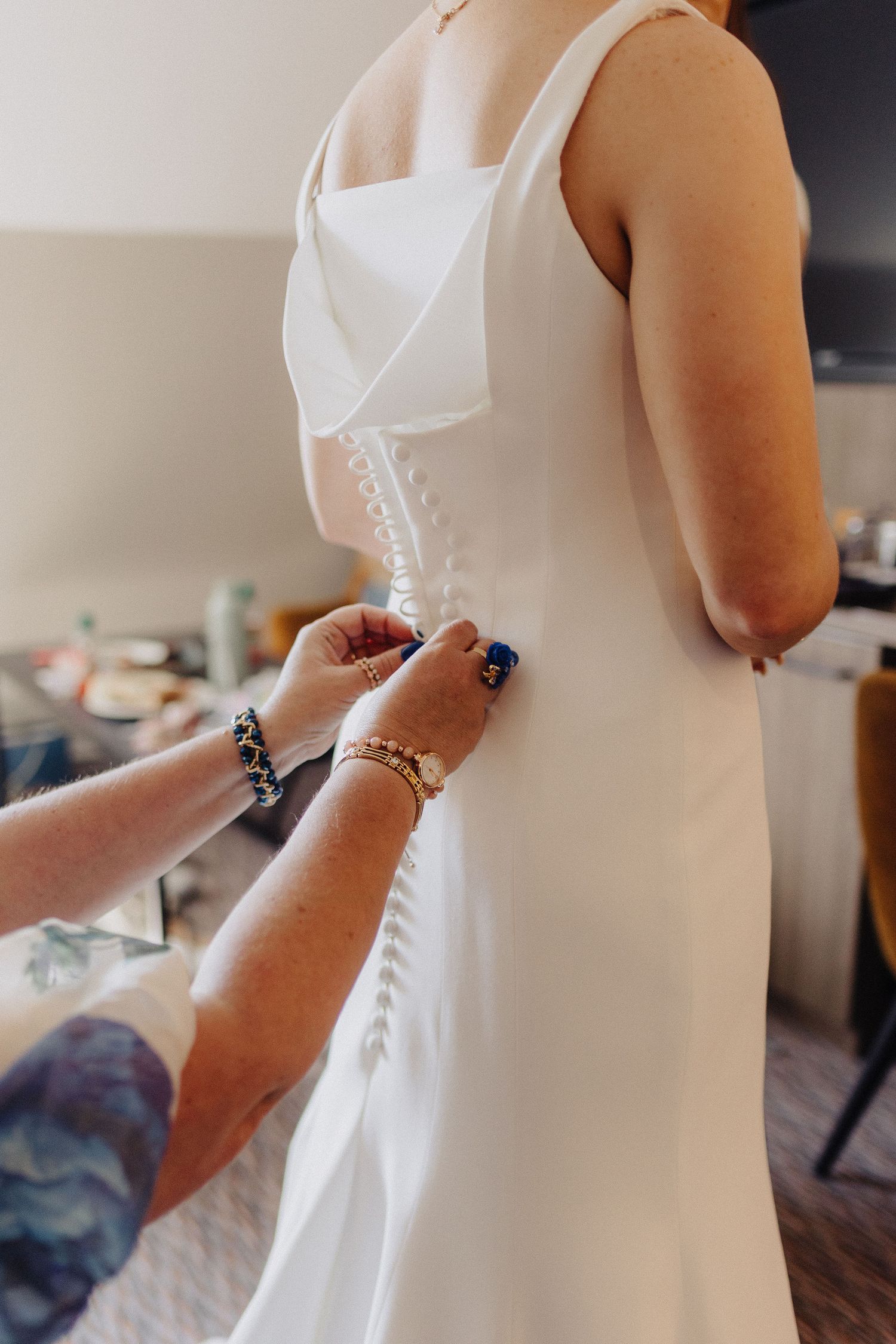 Helper fastening row of pearl buttons on back of brides sleek white wedding dress.