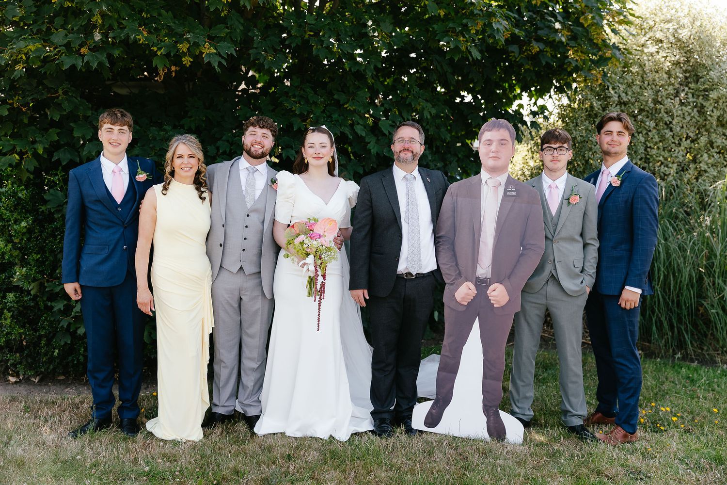 Wedding party posing outdoors with bride and groom surrounded by family members in formal attire before green trees.