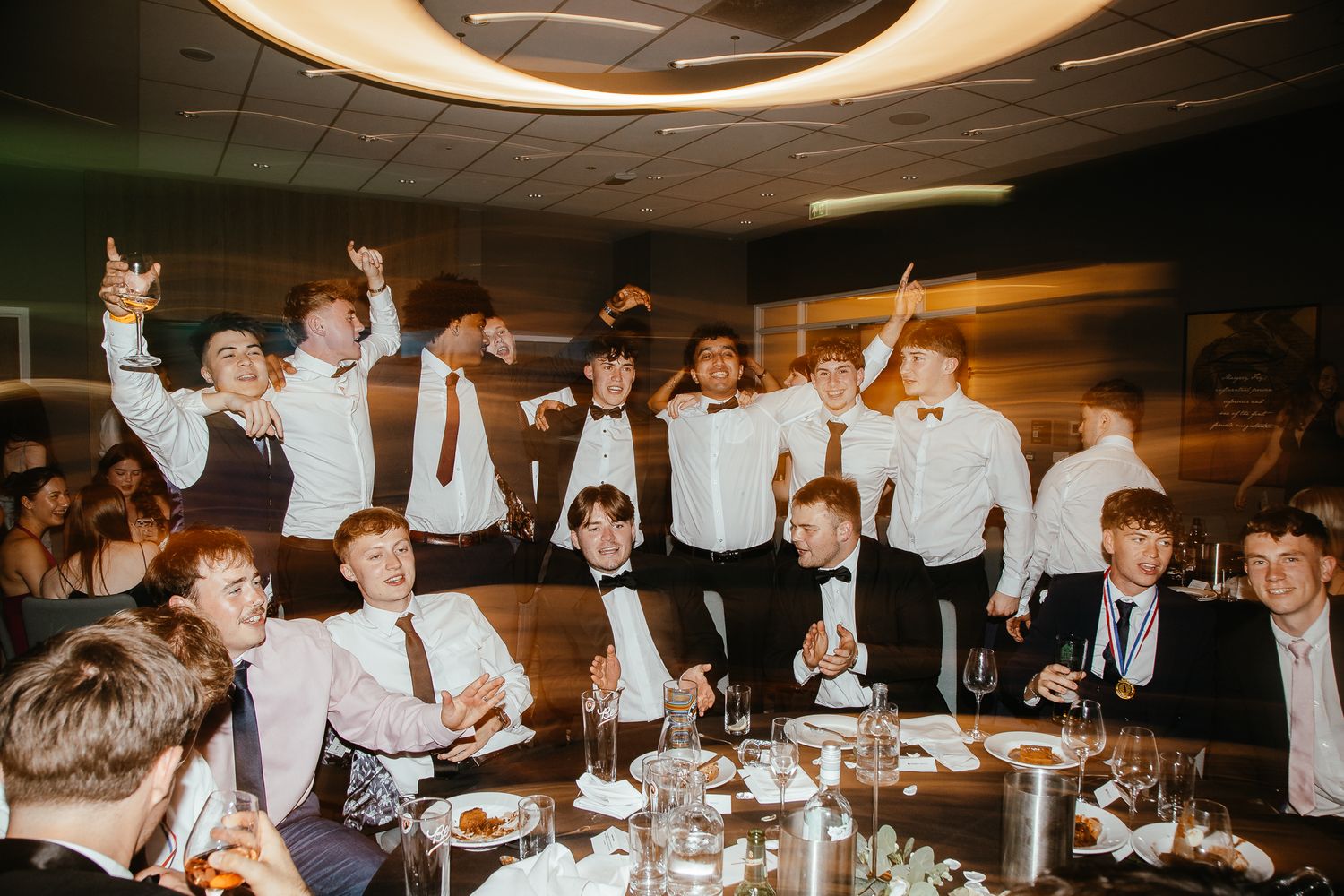 Formal dinner celebration with guests in suits raising their hands enthusiastically.