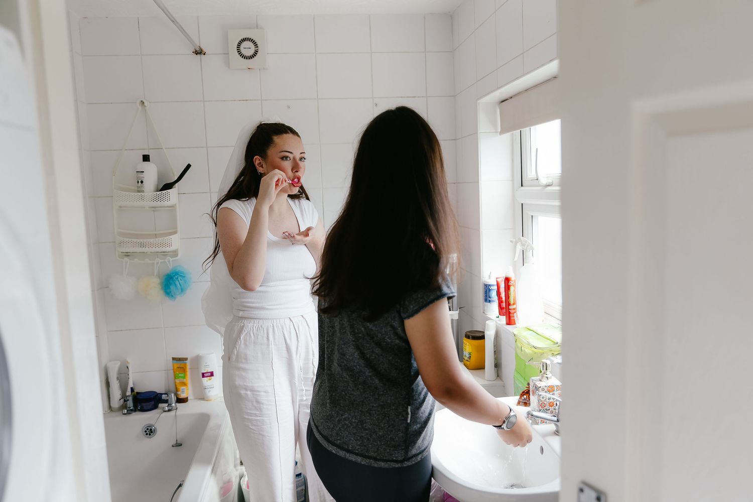 Two women sharing a bright white bathroom, one applying makeup while the other washes hands by the window.