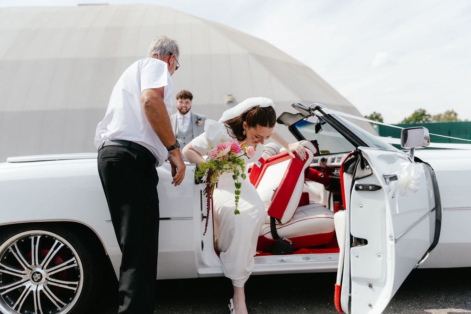 Bride in white dress stepping out of a white convertible limousine with red interior, assisted by a man.