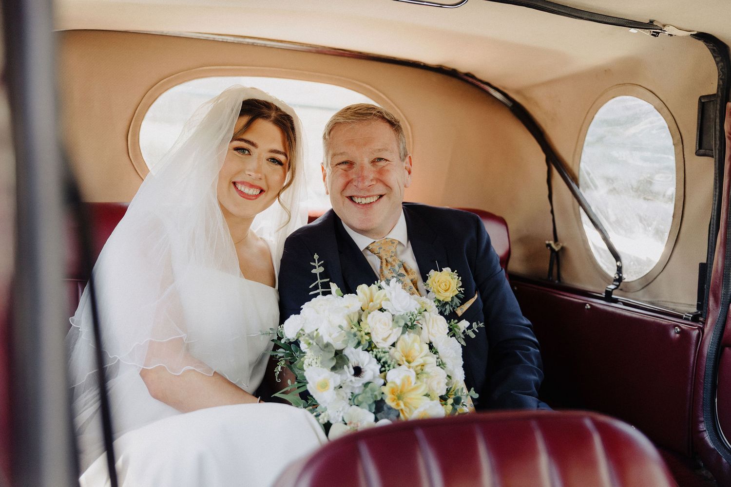 Bride and groom smiling together in a vintage wedding car, bride holding white and yellow bouquet.