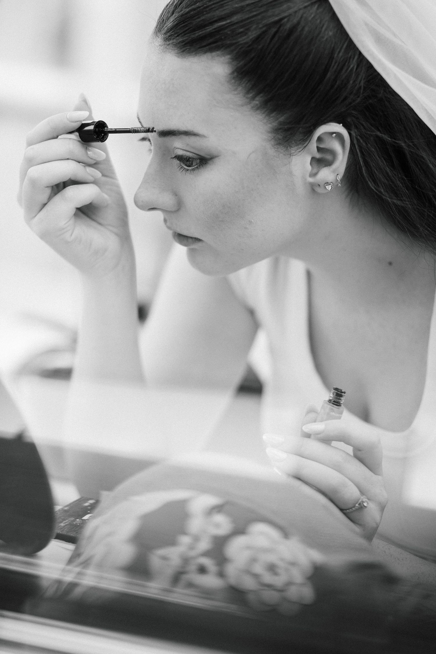 Black and white photo of a bride applying mascara while holding perfume, preparing for her wedding day.