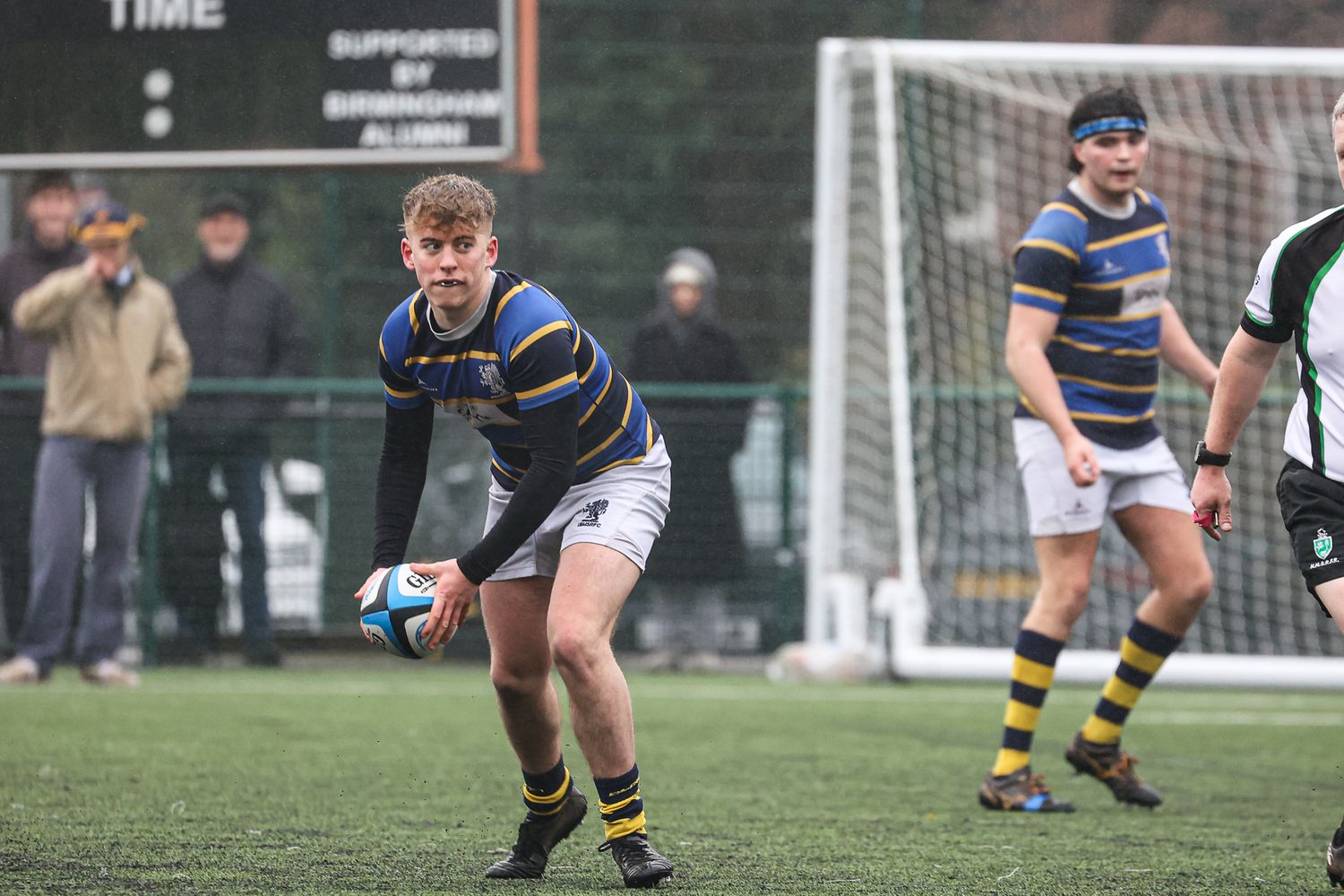 Rugby players sprint across the field during a competitive match with goal posts visible.