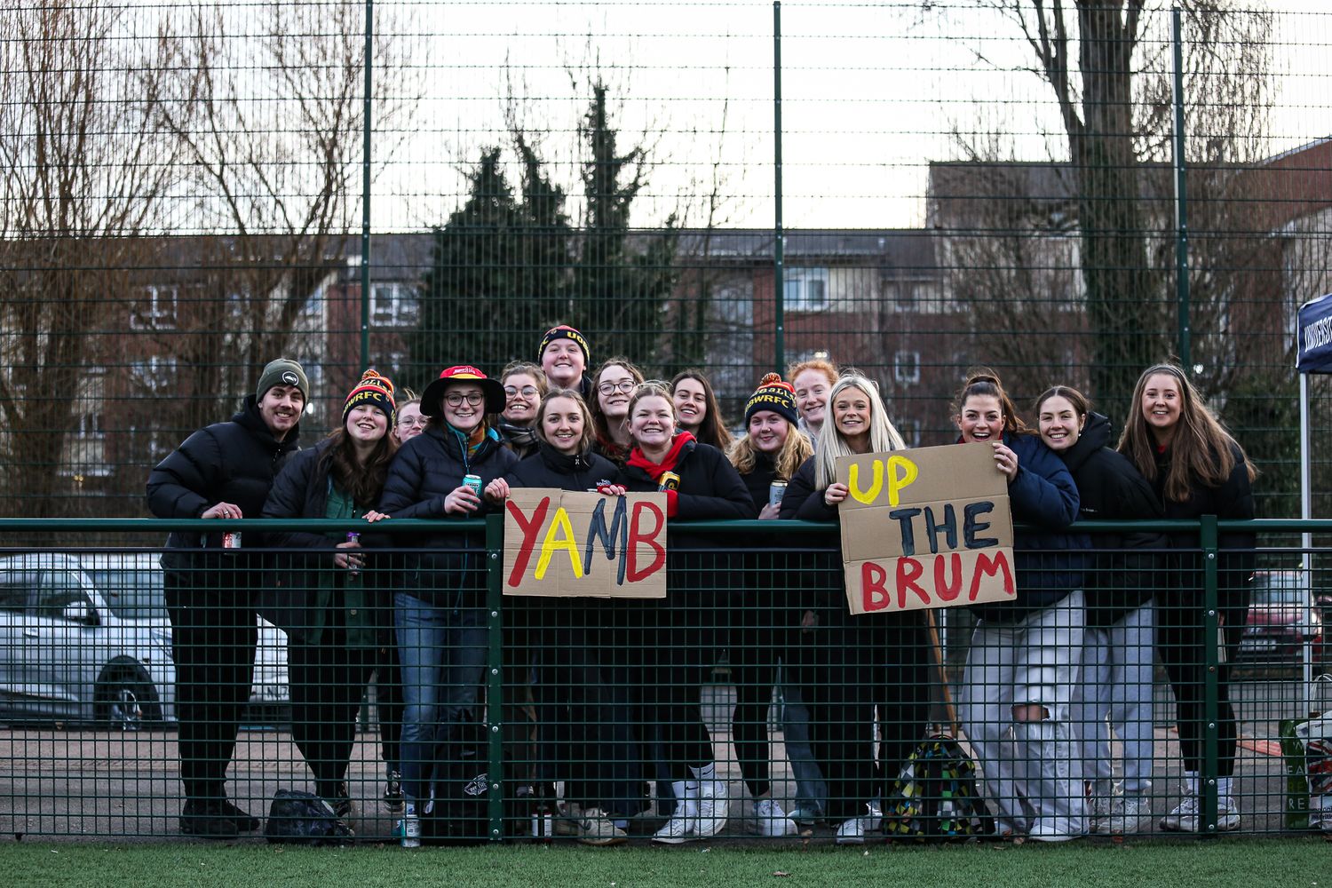 Crowd of supporters holding signs and cheering from behind a fence at a sports match.