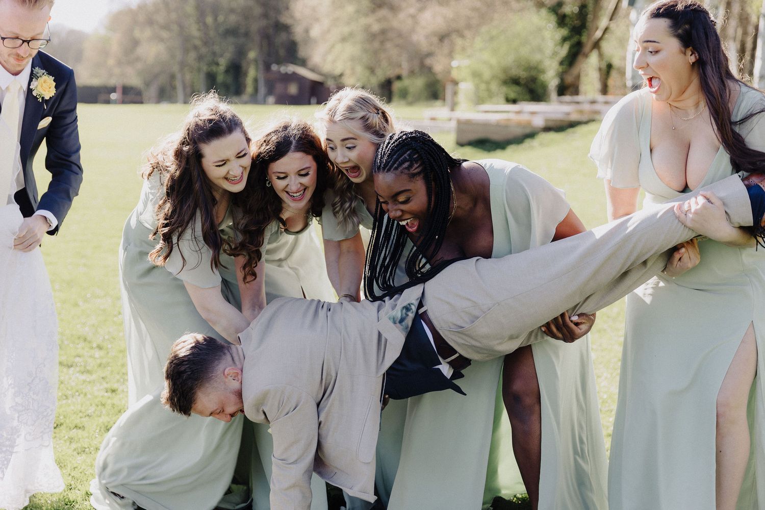Bridesmaids in sage green dresses playfully lifting a groomsman outdoors at a wedding celebration.