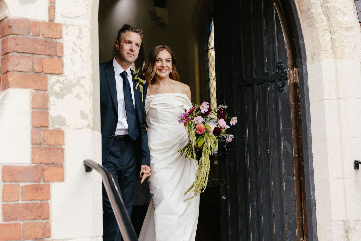 Bride and groom smiling at church doorway, bride in off-shoulder white gown holding colorful floral bouquet.