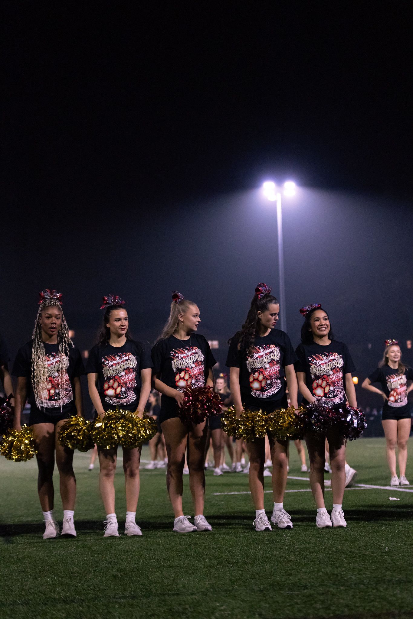 Cheerleaders in pink and black uniforms line up on a football field at night under bright stadium lights.