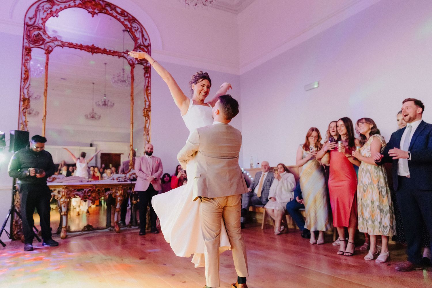 Groom lifting bride during first dance at elegant wedding reception with guests watching and applauding.
