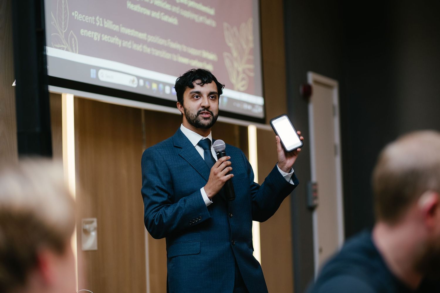 A person in a navy blue suit gestures while speaking at a business presentation in a conference room.