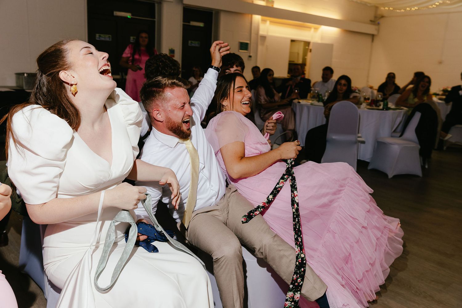 Bride and guests laughing joyfully at a wedding reception, seated together in a decorated event venue.