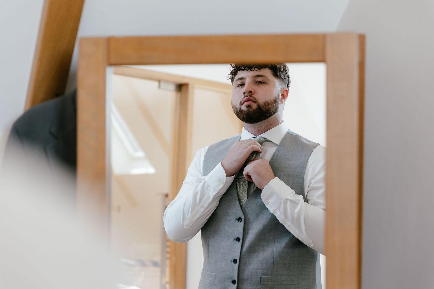 A groom adjusts his tie in a mirror while wearing a grey vest and white dress shirt before his wedding.