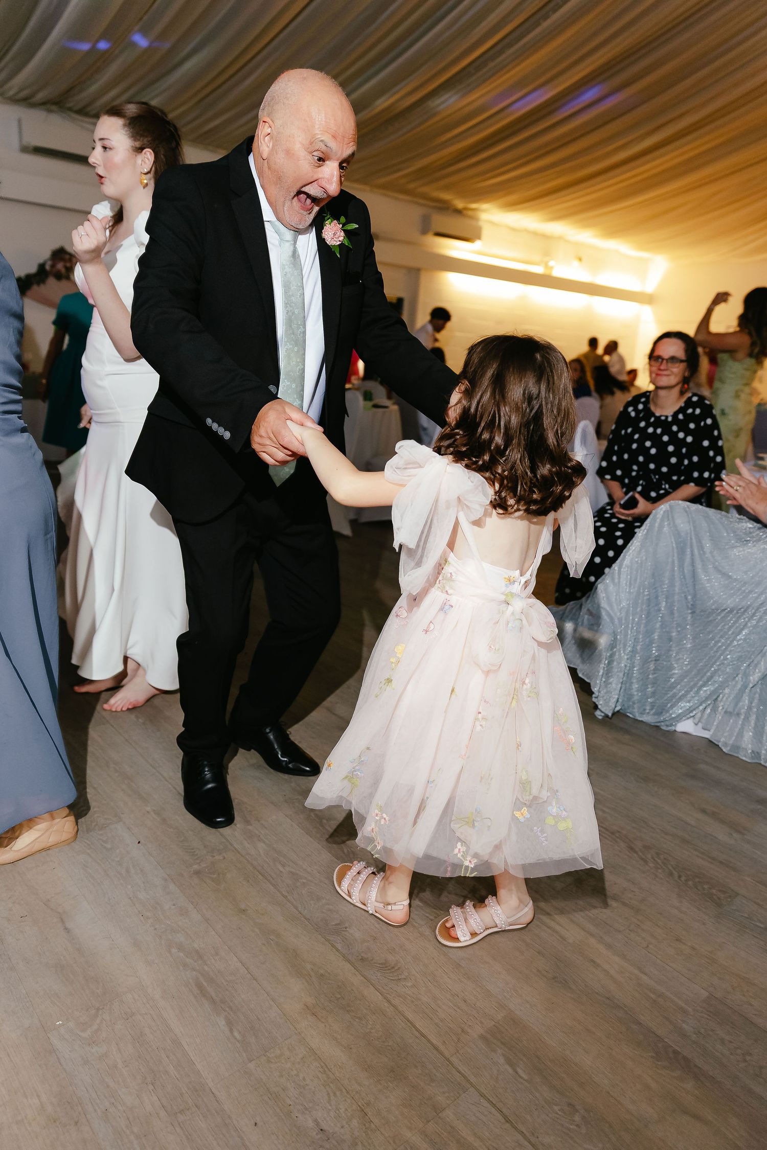 An elderly man in a suit joyfully dances with a young girl in a white dress at a wedding reception.
