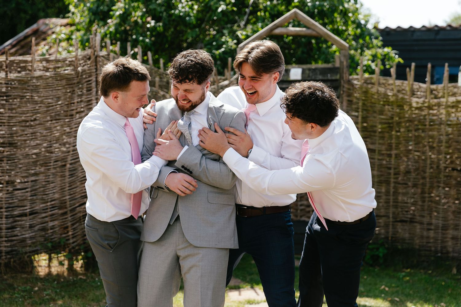 Groomsmen in white shirts and pink ties laughing and adjusting the grooms grey suit jacket outdoors.