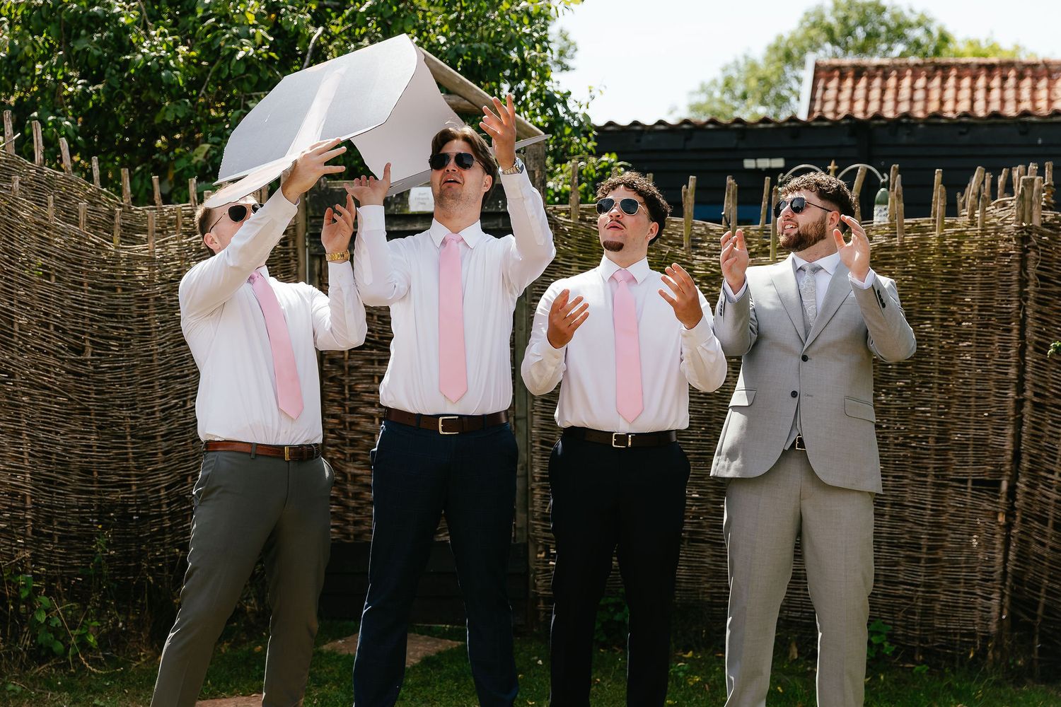 Four groomsmen in pink ties and sunglasses pose outdoors near a wicker fence at a summer wedding.