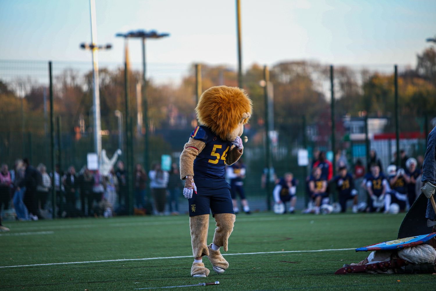 A sports mascot in a navy jersey entertains spectators on an artificial turf field at dusk.