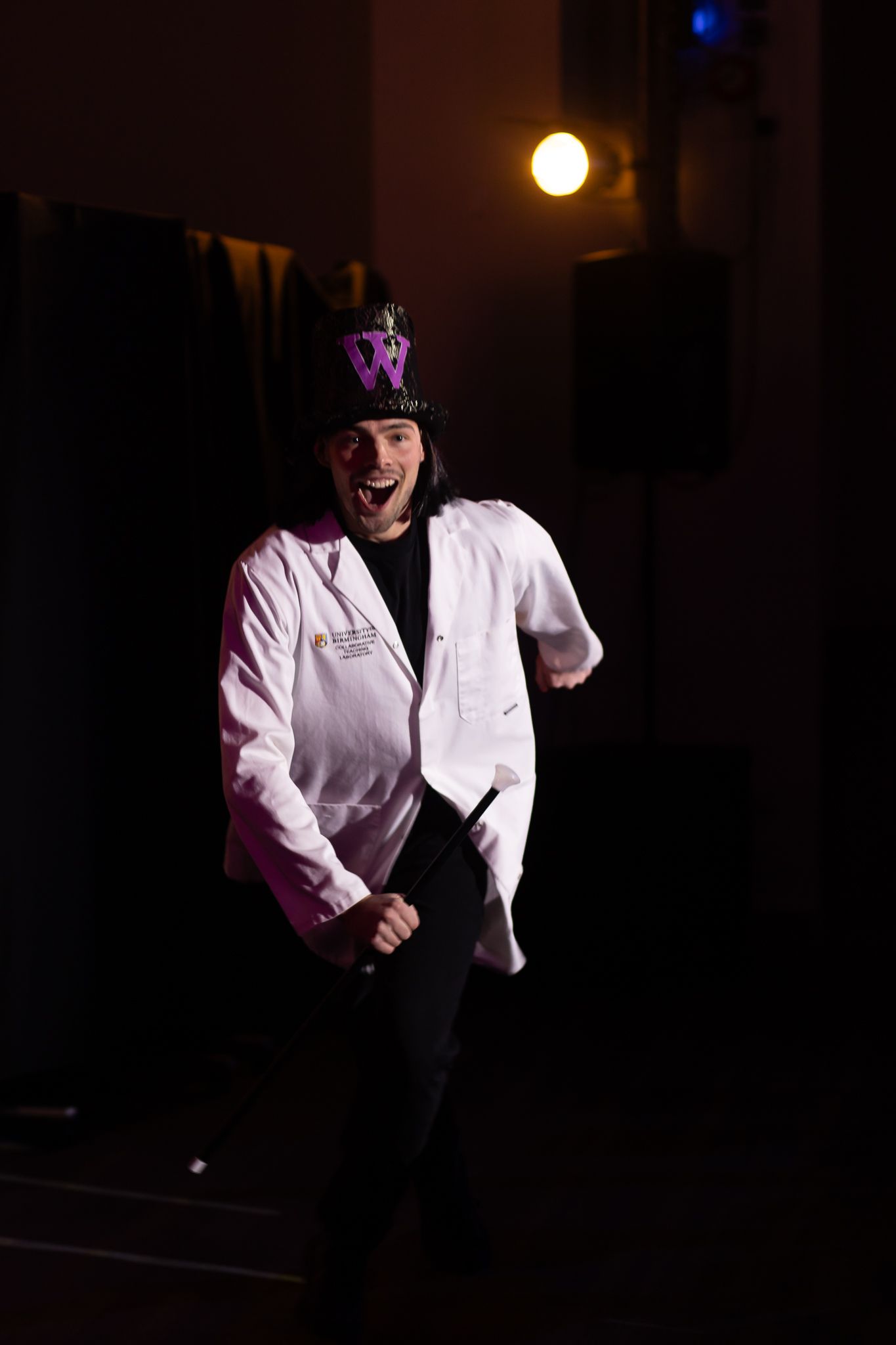 Performer in white lab coat dancing in dramatic lighting on dark stage.