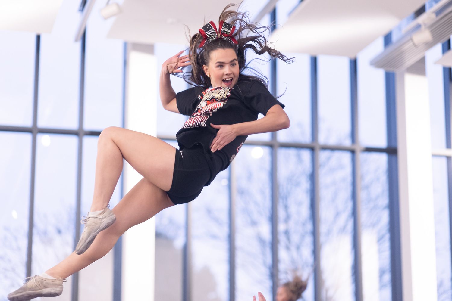 A dancer in a black outfit leaps gracefully through the air in a bright studio space with large windows.