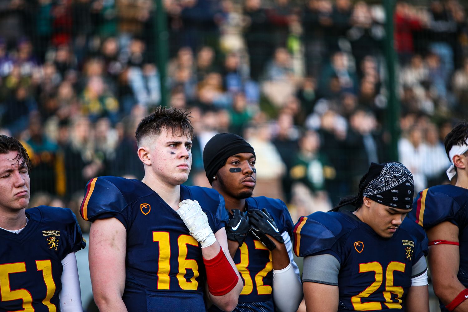Football players in blue and yellow uniforms stand on the sideline during a game.