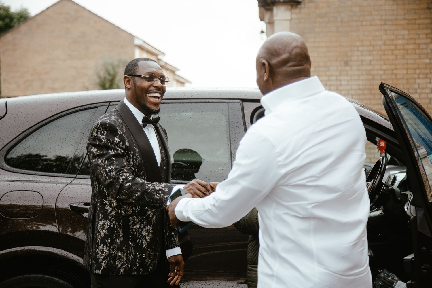 Wedding guests in formal attire greeting each other outside black vehicles.