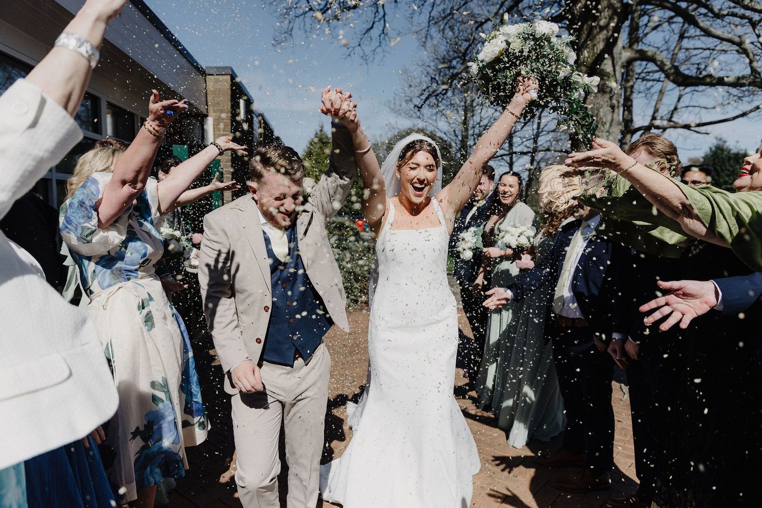 Joyful newlyweds walk through confetti shower surrounded by cheering guests at outdoor wedding celebration.