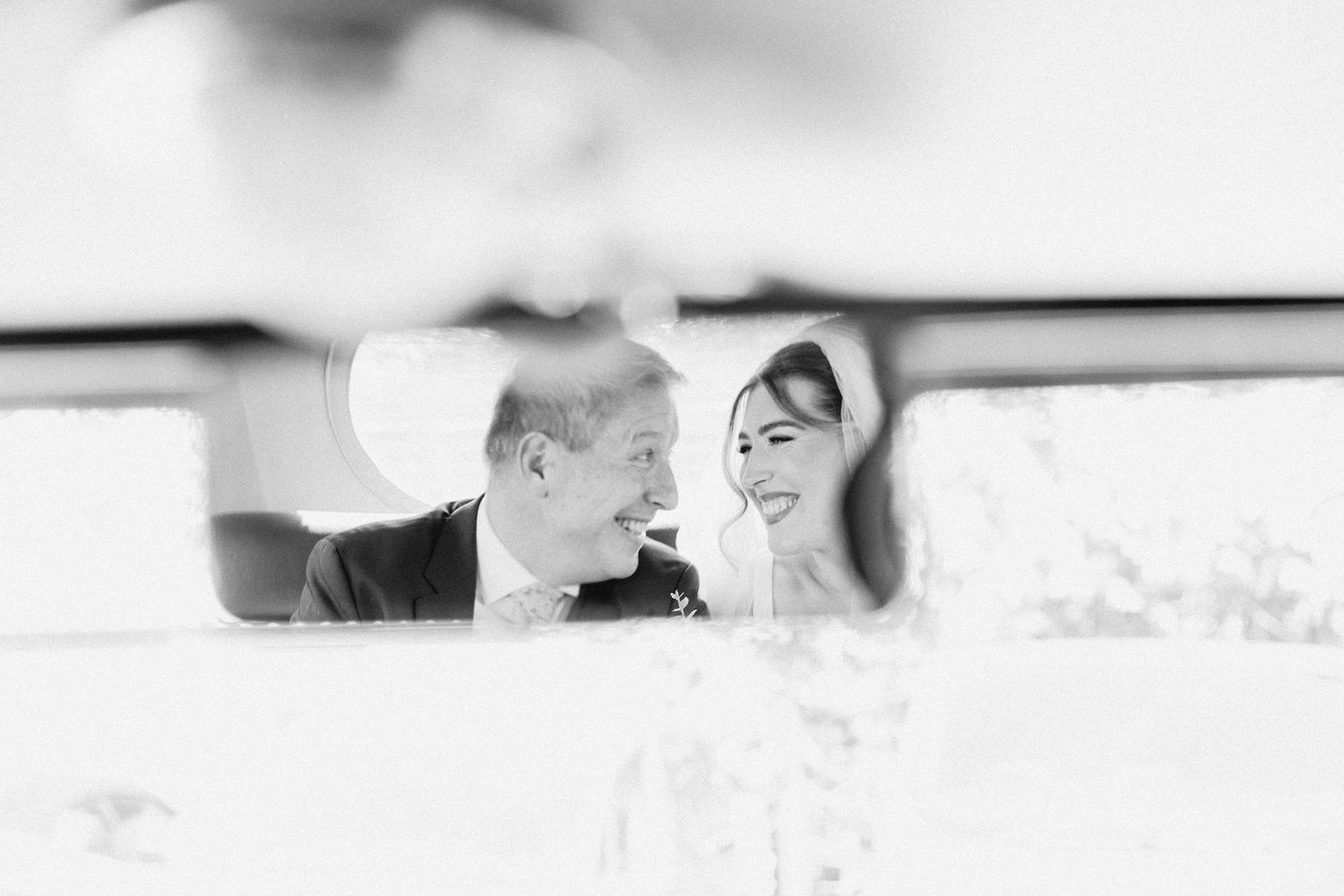 Black and white photo of smiling bride and groom reflected in car mirror.