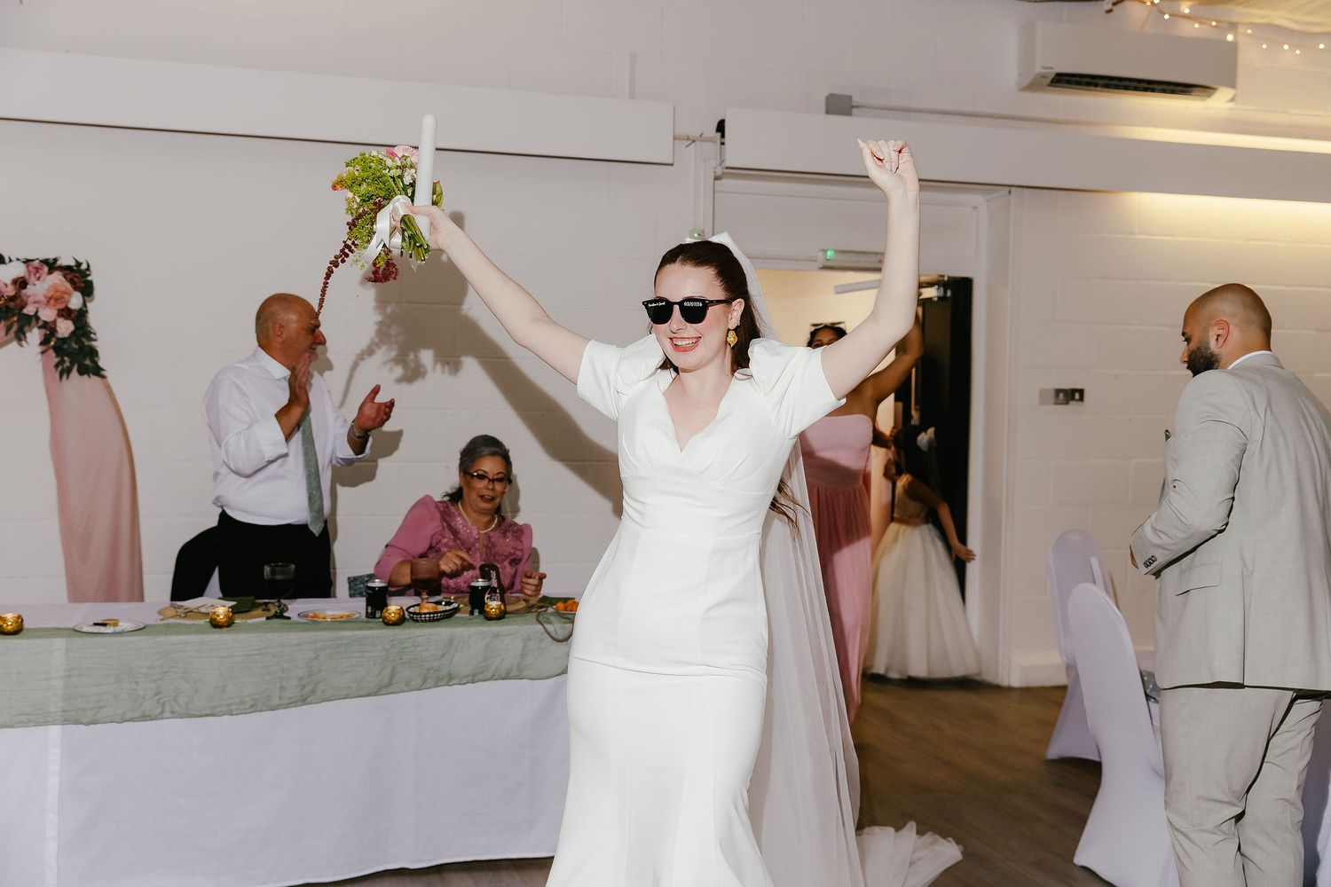 Bride in white dress and sunglasses celebrates at wedding reception, raising bouquet with arms up while guests applaud.