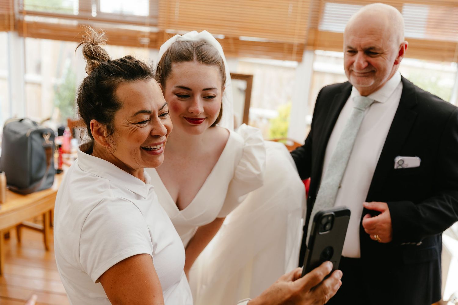 Bride in white gown smiling with guests viewing photos on a smartphone before the wedding ceremony.