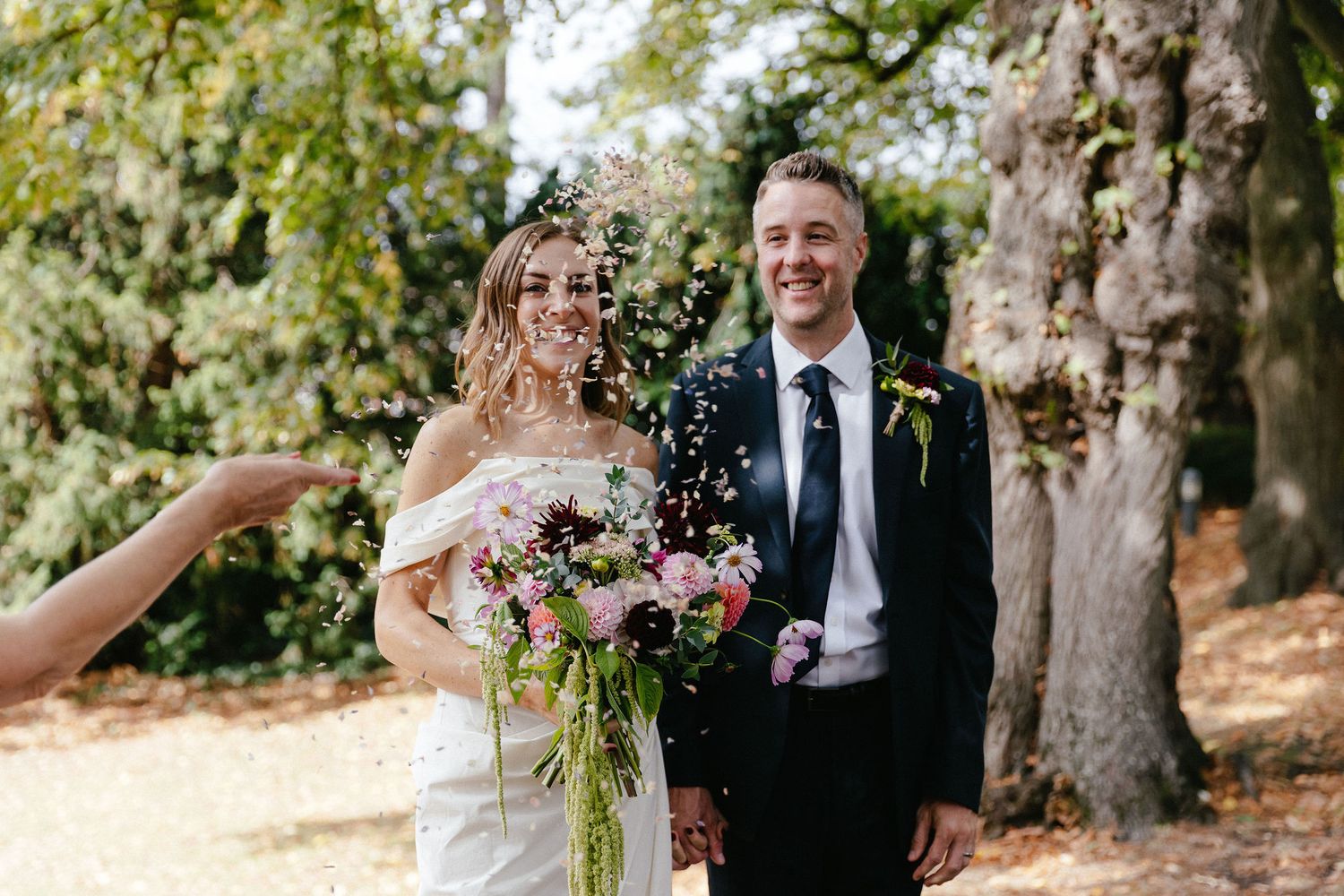 Bride and groom smiling outdoors as confetti is thrown, bride holding colorful bouquet, groom in black suit with tie.