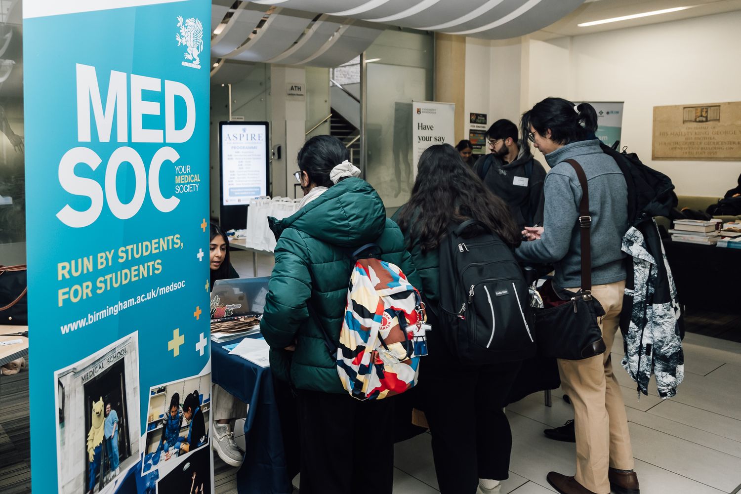 Students line up at a MedSoc information booth displaying a blue banner and promotional materials.